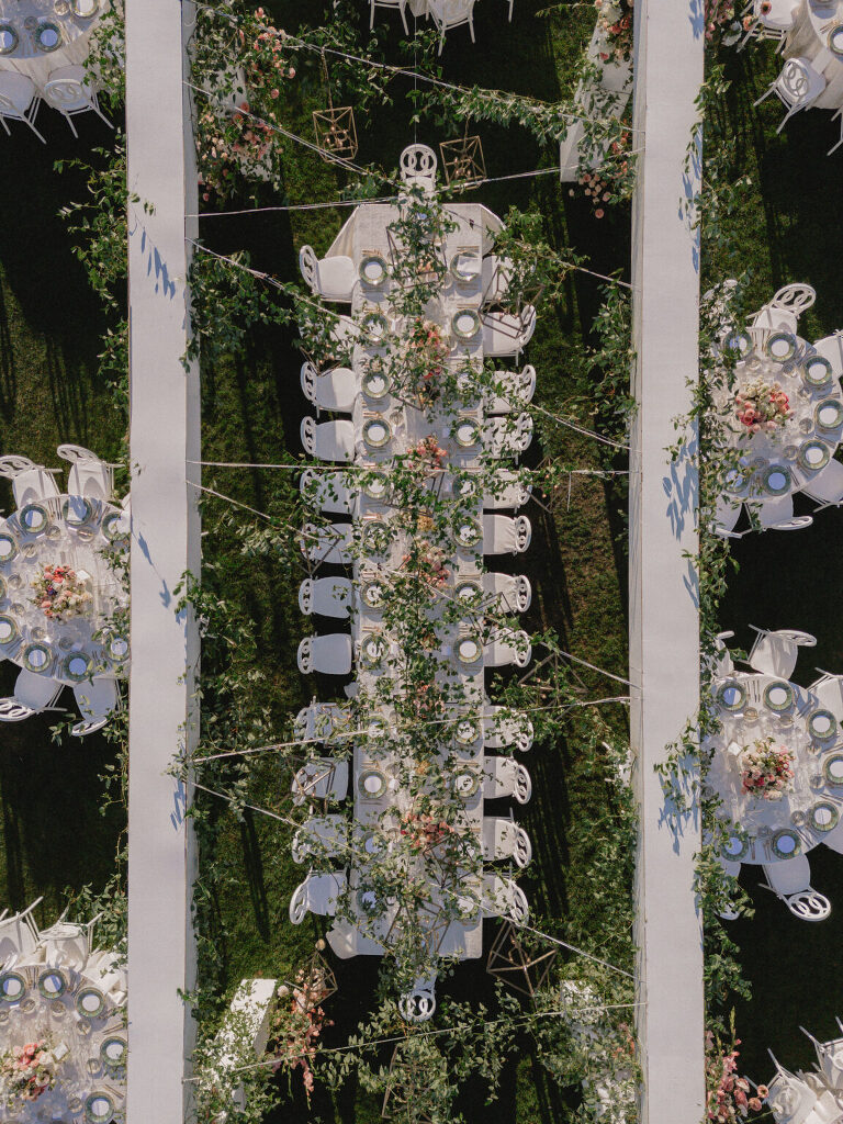 A garden reception seen straight down: a long table wrapped in greenery, round tables and lanterns across the lawn