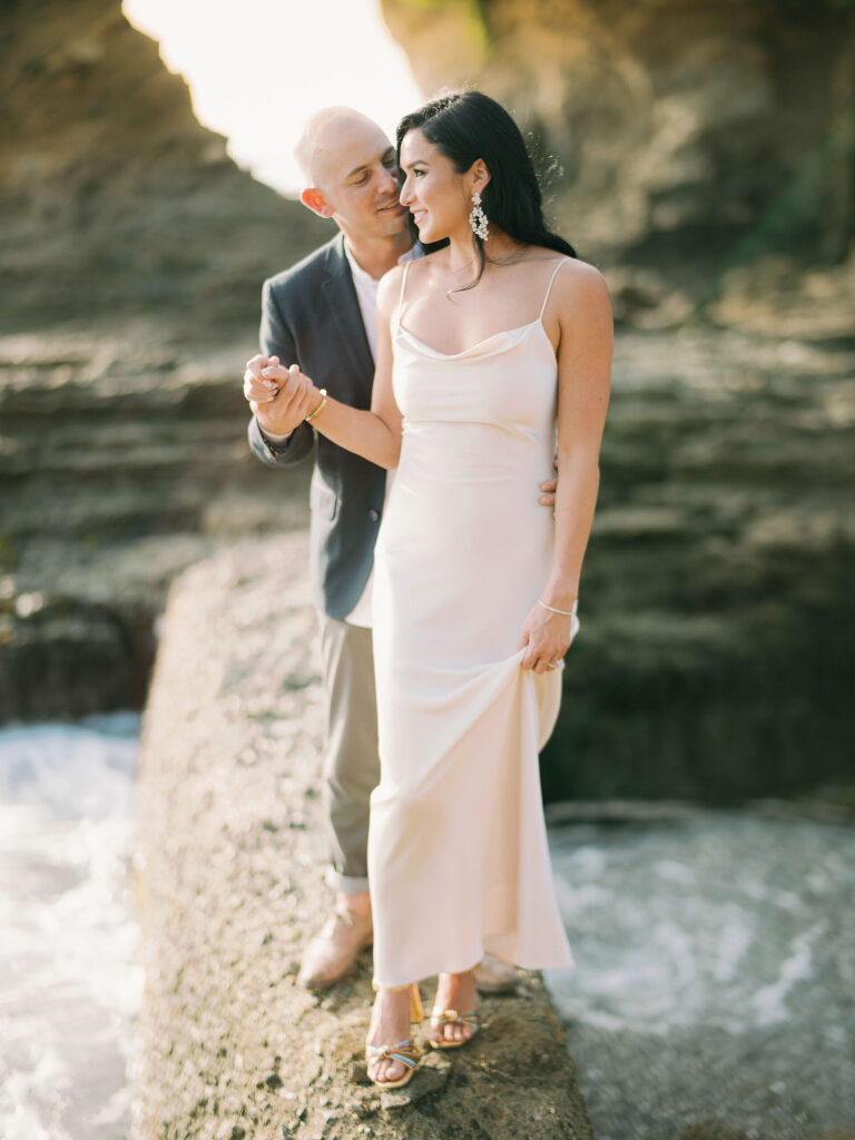 A couple on wet rocks in a golden backlit cove, surf washing behind them
