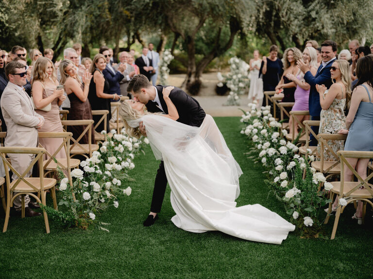 The groom dips and kisses the bride mid-aisle while guests stand and applaud in an olive grove