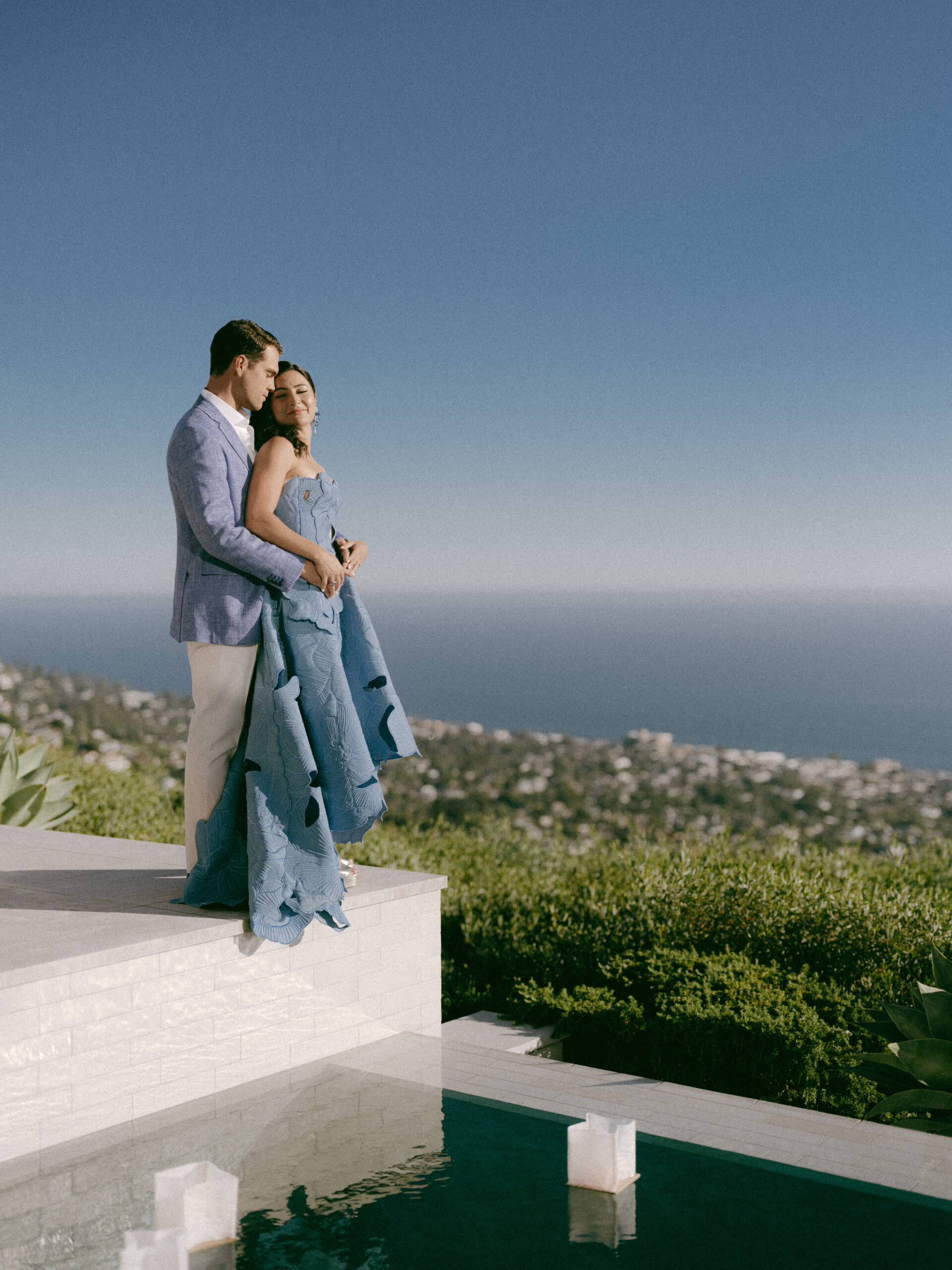 A couple embracing on a white terrace above a pool of floating candle lanterns at dusk