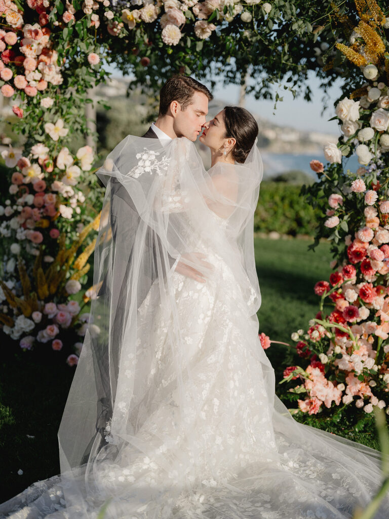 Bride and groom nose to nose beneath a floral arch, the Pacific coastline glowing behind them