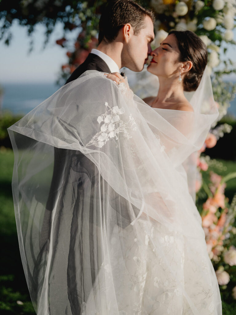 The couple kissing at the floral arch, wind catching the embroidered veil