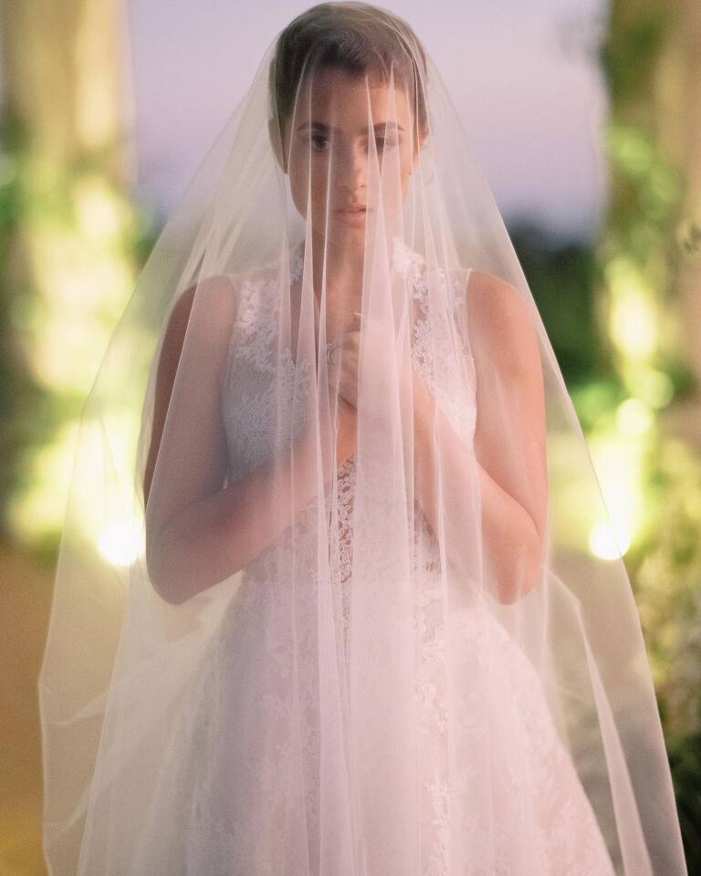 The bride beneath her veil at dusk in a garden colonnade, hands gathered, lights warm behind her