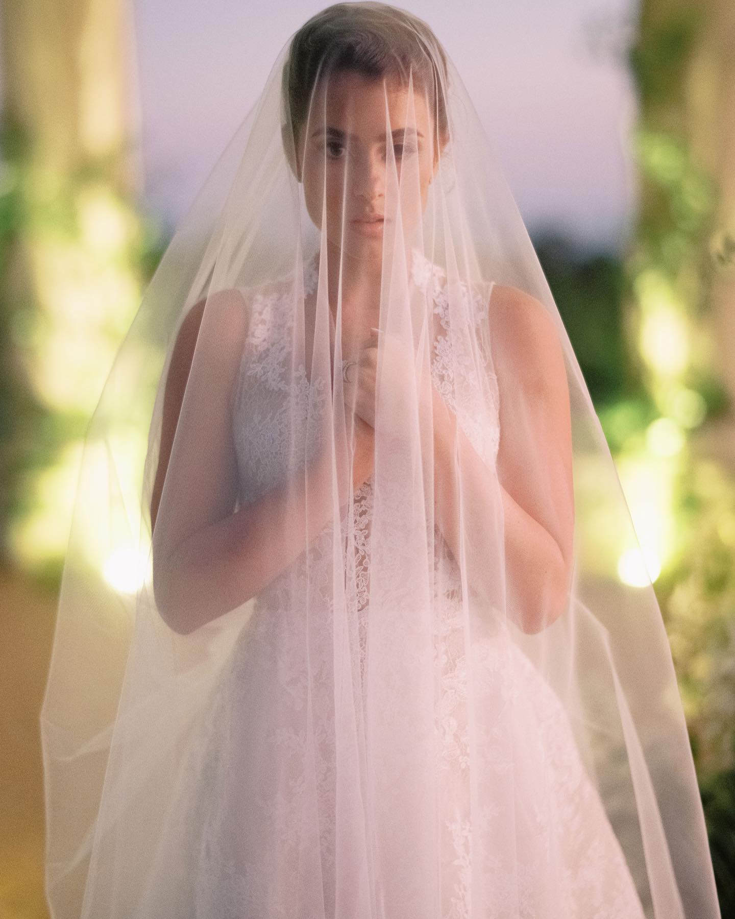 The bride beneath her veil at dusk in a garden colonnade, hands gathered, lights warm behind her