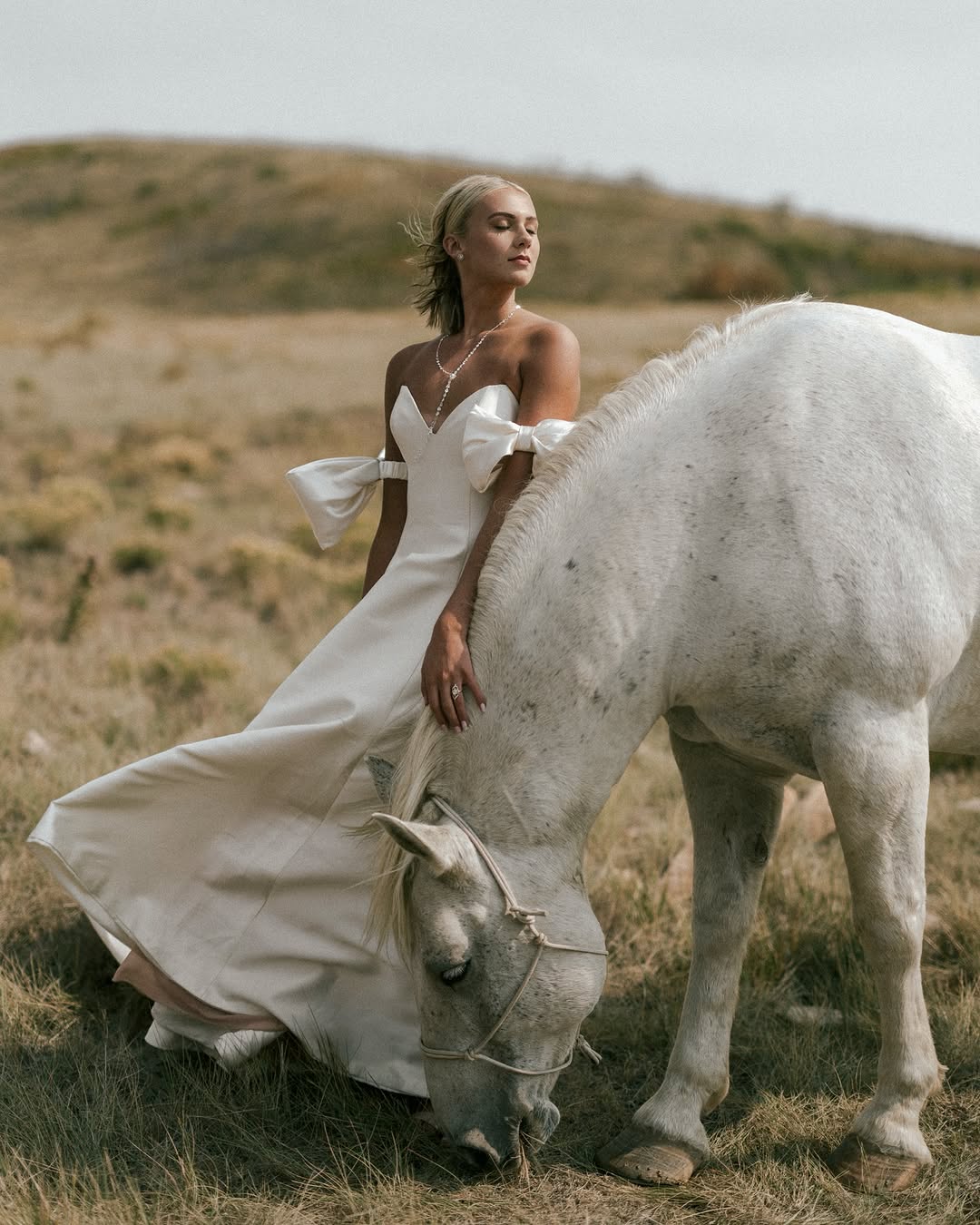 A bride in a bow detail satin gown beside a grazing white horse in golden grass hills