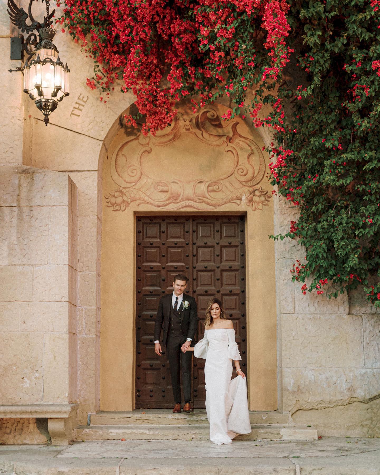 The couple stepping from a studded wooden doorway beneath cascading bougainvillea, a lantern lit beside them