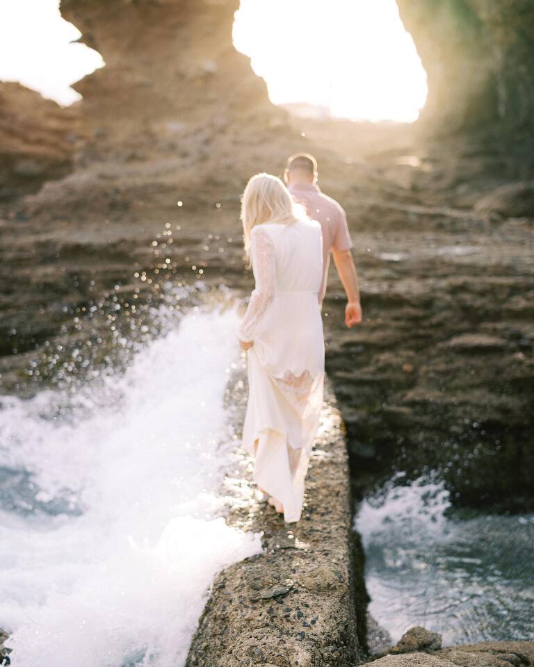 The couple walking a rock ledge through a sea cave, surf breaking beside them in golden light