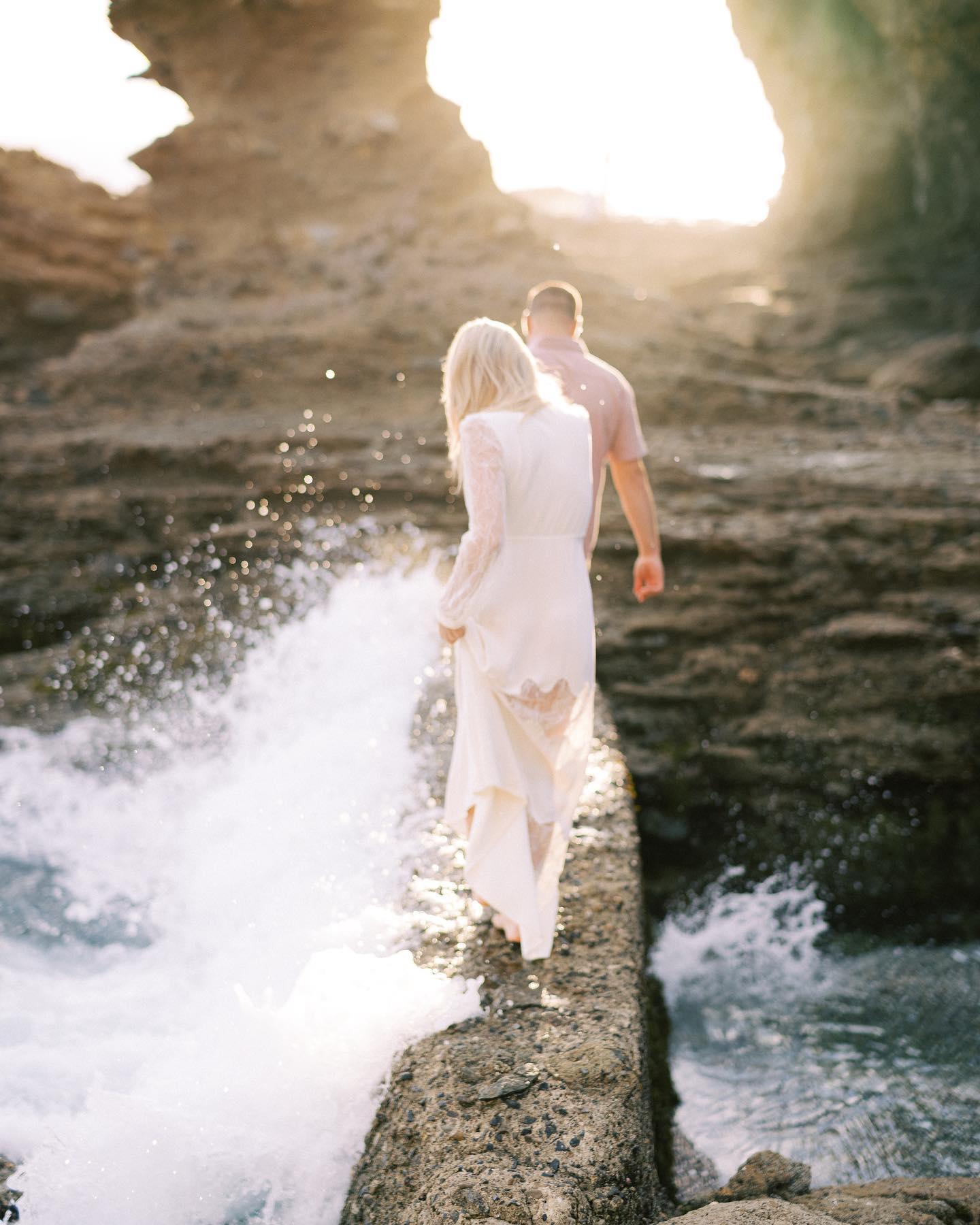 The couple walking a rock ledge through a sea cave, surf breaking beside them in golden light