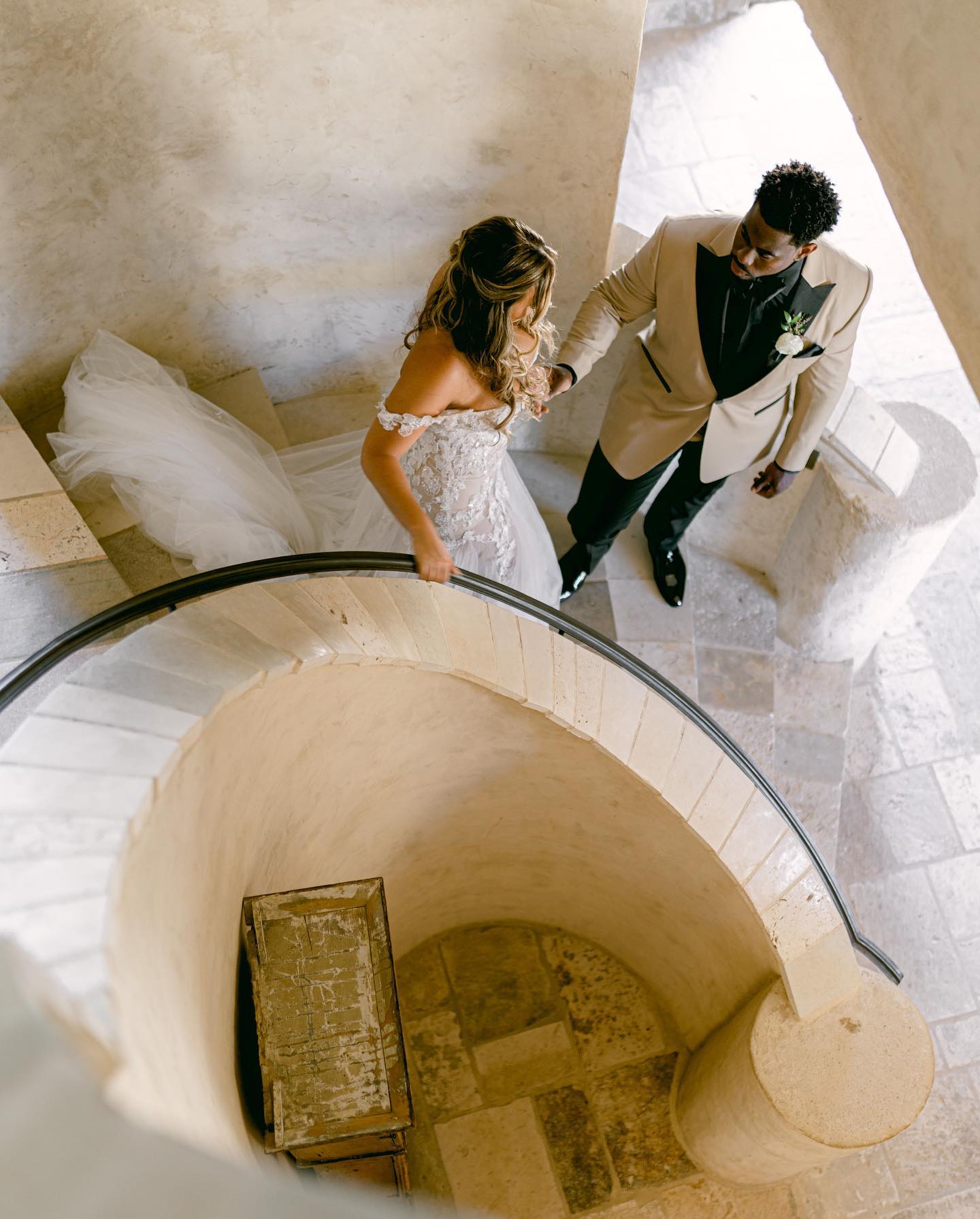 The couple descending a limestone spiral staircase seen from above, the veil trailing on the steps