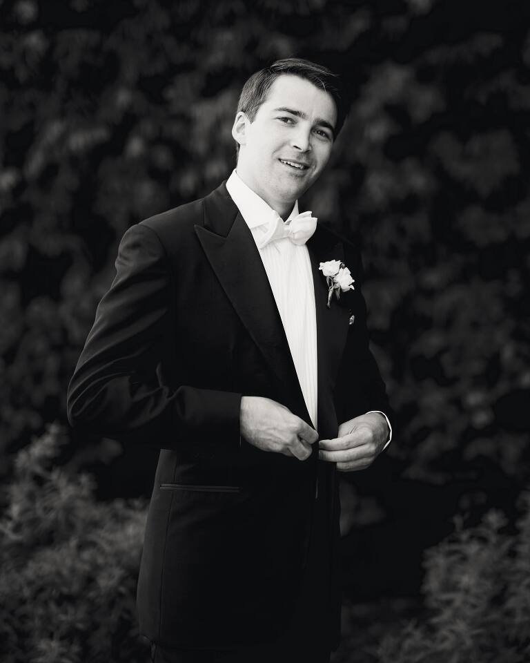 A monochrome portrait of the groom in white tie, fastening his jacket before the ceremony